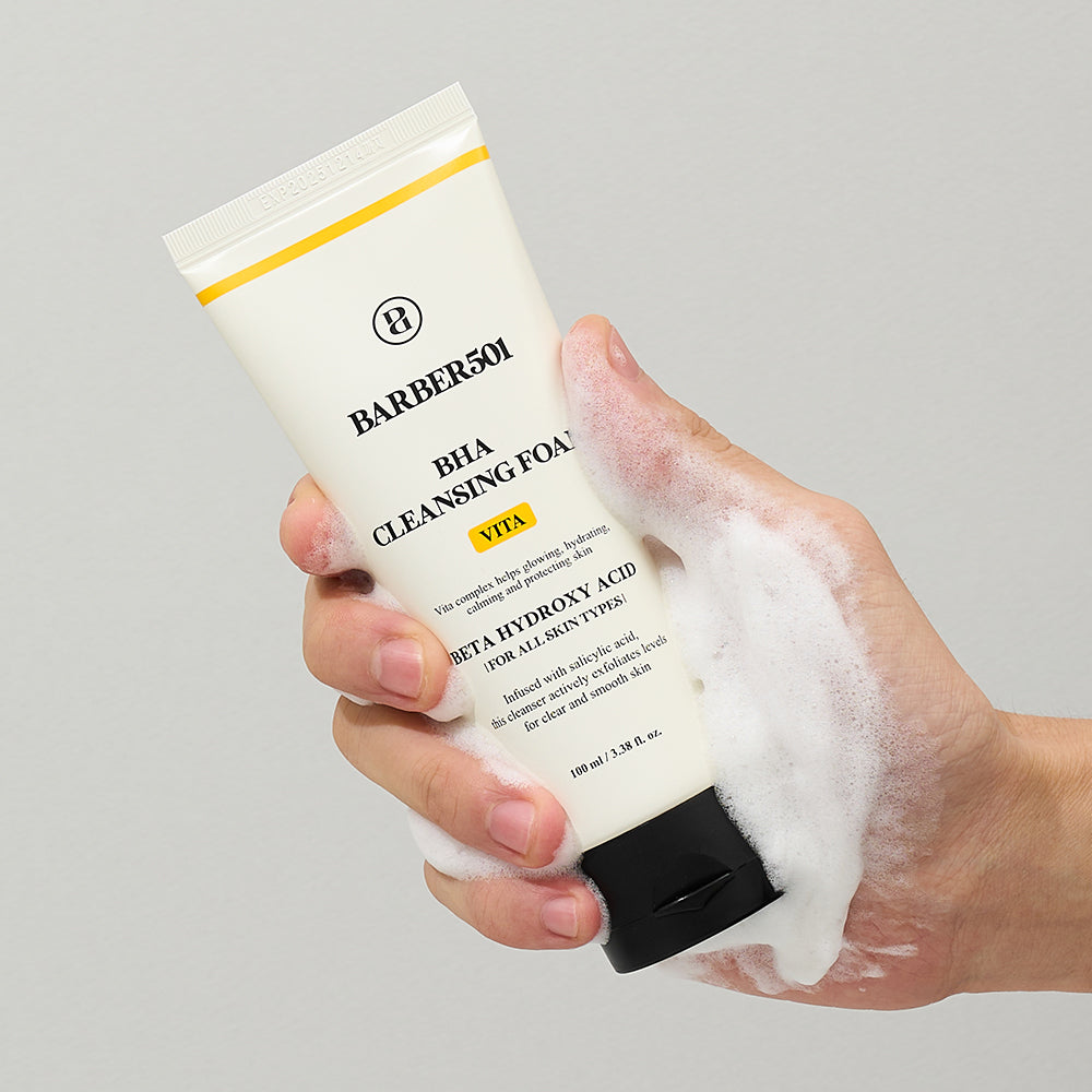 Hand holding a tube of BARBER501 BHA Cleansing Foam with foam on a gray background