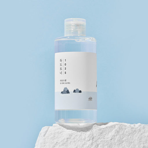 A clear plastic bottle containing a liquid toner with a label that has text and a graphic design, placed on a white stone surface with a blue background.