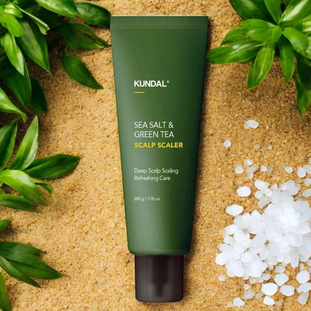 A tube of KUNDAL Sea Salt & Green Tea Scalp Scaler resting on a sandy background, surrounded by green leaves and white salt crystals.