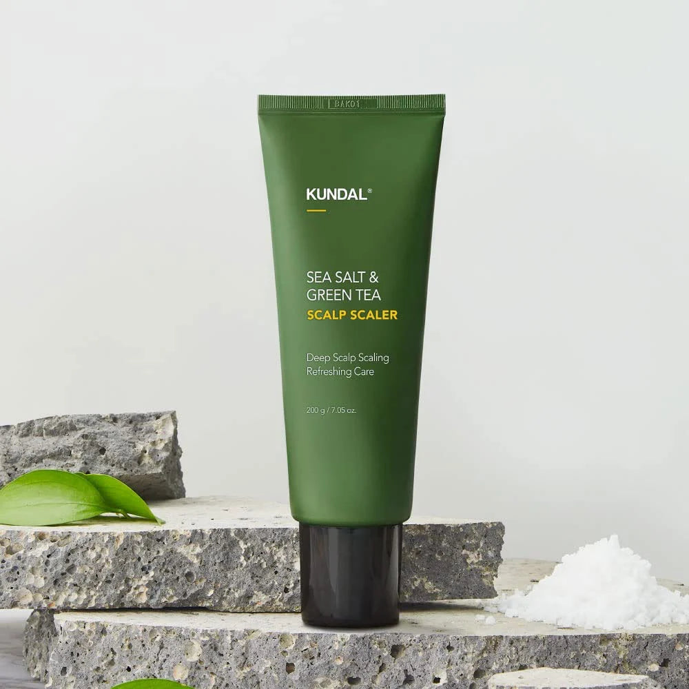A tube of KUNDAL Sea Salt & Green Tea Scalp Scaler standing upright on stacked gray stone slabs, with a small pile of white salt and green leaves nearby on a light neutral background.