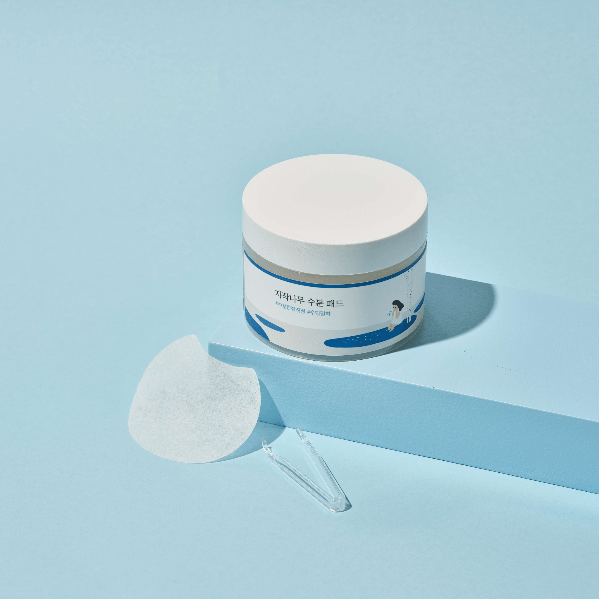 A white jar with a lid containing birch moisturizing pads on a light blue background, with a few pads displayed outside the jar.