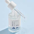 A clear glass bottle with a white dropper containing a gel-like skincare product, labeled 'Round Lab 1025 Dokdo Ampoule 45g'.