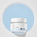 A white jar of Round Lab 1025 DOKDO Cream with a label that includes a blue and green graphic, possibly representing skin or nature.
