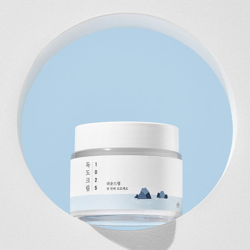 A white jar of Round Lab 1025 DOKDO Cream with a label that includes a blue and green graphic, possibly representing skin or nature.